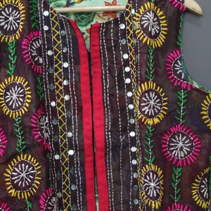 Puede incluir: Prenda reversible sin mangas con estampado floral. La capa exterior es marr&oacute;n con flores bordadas en amarillo, rosa y blanco. La capa interior tiene un estampado floral verde claro. Una franja roja recorre el centro.