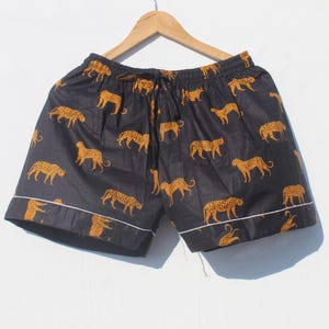 Può includere: Pantaloncini neri con un motivo ripetuto di ghepardi arancioni. I pantaloncini hanno un girovita con coulisse e bordi bianchi. Sono appesi a una gruccia di legno su sfondo bianco.