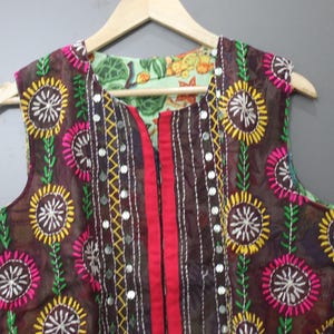 Puede incluir: Chaleco sin mangas marr&oacute;n con bordados florales coloridos. El chaleco presenta un panel central rojo y est&aacute; adornado con peque&ntilde;os espejos. El forro es de tela verde claro con un estampado floral. El chaleco est&aacute; colgado en una percha de madera.