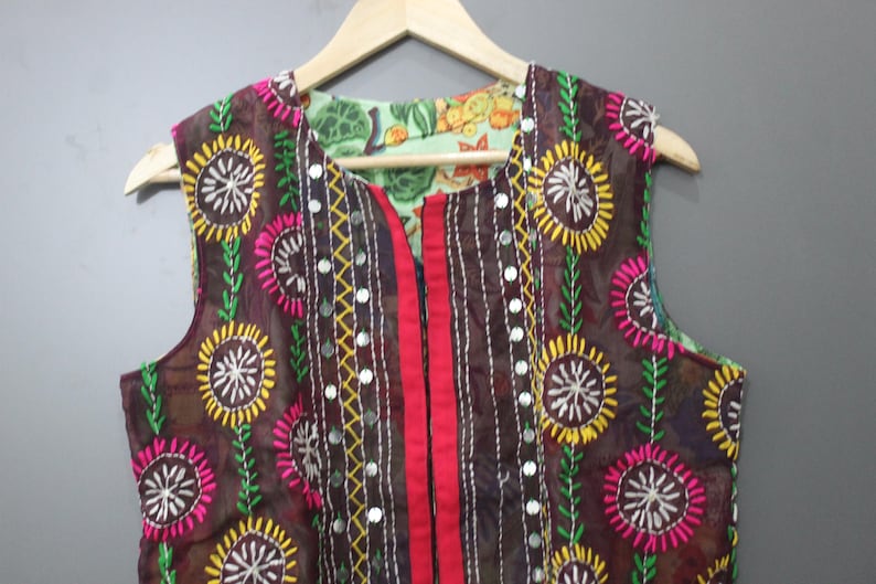 Puede incluir: Chaleco sin mangas marr&oacute;n con bordados florales coloridos. El chaleco presenta un panel central rojo y est&aacute; adornado con peque&ntilde;os espejos. El forro es de tela verde claro con un estampado floral. El chaleco est&aacute; colgado en una percha de madera.