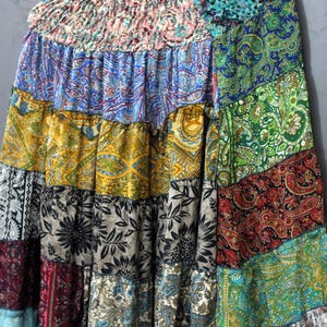 May include: A patchwork skirt with a variety of colourful patterns, including paisley, floral, and geometric designs. The skirt is made of multiple fabric panels in shades of blue, green, yellow, red, and black. The skirt is hanging on a wooden hanger.