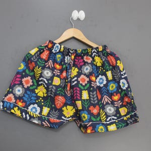 May include: Navy blue shorts featuring a vibrant, multi-colored floral and botanical pattern in shades of yellow, red, light blue, pink, and white. The shorts have an elastic waistband with a drawstring and white piping along the bottom hem.
