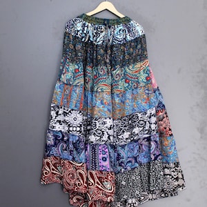 May include: A long, patchwork skirt with a variety of paisley and floral patterns in shades of blue, red, purple, and black. The skirt is hanging on a wooden hanger against a gray background.