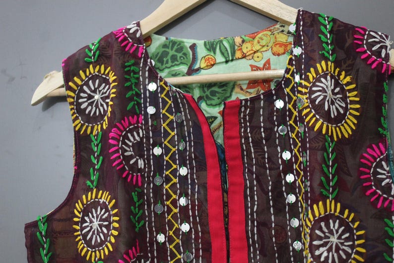 Puede incluir: Un chaleco marr&oacute;n sin mangas con bordados coloridos. El chaleco presenta dise&ntilde;os florales amarillos, rosas y blancos, detalles de hojas verdes y peque&ntilde;os adornos plateados. El chaleco es reversible, con un forro estampado floral.