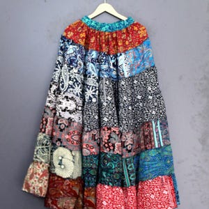 Könnte beinhalten: Langer Patchwork-Rock mit einer Vielzahl farbenfroher Blumen- und Paisley-Muster. Der Rock besteht aus mehreren Stoffbahnen in Rot-, Blau-, Grün- und Schwarztönen. Er hängt an einem Holzbügel vor grauem Hintergrund.