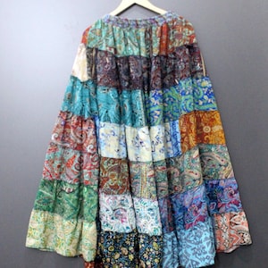 Könnte beinhalten: Ein Patchwork-Rock mit Stufen-Design, der eine Vielzahl farbenfroher Stoffe mit Mustern aufweist. Der Rock wird auf einem Holzbügel vor grauem Hintergrund präsentiert. Die Stoffe umfassen Paisley-, Blumen- und geometrische Muster in Blau-, Grün-, Rot- und Orangetönen.