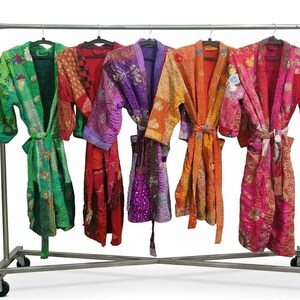 Könnte beinhalten: Fünf langärmelige Morgenmäntel mit Bindegürteln hängen an einer Kleiderstange aus Metall. Die Mäntel weisen Patchwork-Designs in leuchtenden Farben wie Grün, Rot, Violett, Orange und Pink auf, mit durchgehenden, feinen Steppnähten.