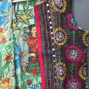 Puede incluir: Un chaleco sin mangas con un dise&ntilde;o colorido y en capas. El chaleco presenta un estampado floral y bot&aacute;nico en el interior, un borde rojo y una capa exterior marr&oacute;n oscuro con patrones florales bordados y detalles de espejo. El chaleco est&aacute; colgado de una percha de madera.