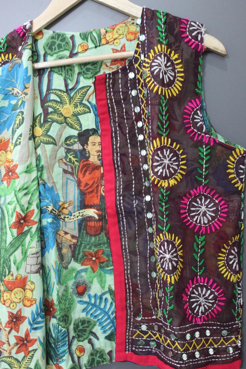 Puede incluir: Un chaleco sin mangas con un dise&ntilde;o colorido y en capas. El chaleco presenta un estampado floral y bot&aacute;nico en el interior, un borde rojo y una capa exterior marr&oacute;n oscuro con patrones florales bordados y detalles de espejo. El chaleco est&aacute; colgado de una percha de madera.