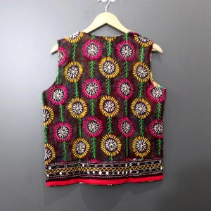 Puede incluir: Un chaleco sin mangas de tejido transparente marr&oacute;n oscuro, adornado con motivos florales bordados en amarillo, rosa y blanco. El chaleco tiene un borde rojo en la parte inferior, adornado con peque&ntilde;os espejos y un ribete decorativo.