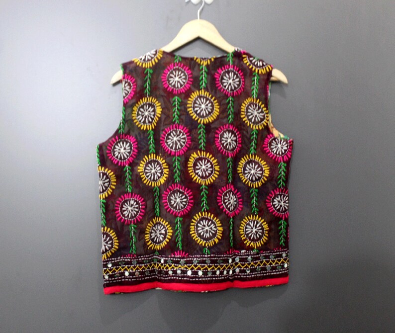 Puede incluir: Un chaleco sin mangas de tejido transparente marr&oacute;n oscuro, adornado con motivos florales bordados en amarillo, rosa y blanco. El chaleco tiene un borde rojo en la parte inferior, adornado con peque&ntilde;os espejos y un ribete decorativo.