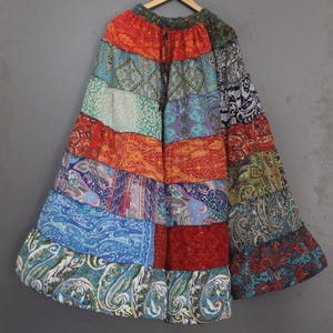 May include: A patchwork skirt with a vibrant array of colors and patterns. The skirt features a mix of paisley, floral, and geometric designs in shades of red, orange, blue, green, and purple. The skirt is hanging on a wooden hanger against a gray background.