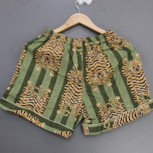 May include: A pair of green shorts featuring a repeating pattern of tigers and vertical stripes. The shorts have an elastic waistband and white piping along the bottom hem, hanging from a wooden hanger against a plain gray background.