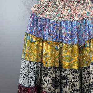 May include: A patchwork skirt with a variety of colourful patterns, including paisley and floral designs. The skirt is made of multiple fabric panels in shades of blue, yellow, black, and red. The skirt is hanging on a wooden hanger.