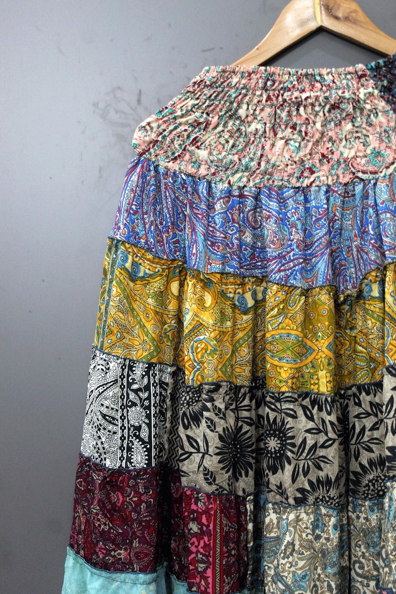 May include: A patchwork skirt with a variety of colourful patterns, including paisley and floral designs. The skirt is made of multiple fabric panels in shades of blue, yellow, black, and red. The skirt is hanging on a wooden hanger.