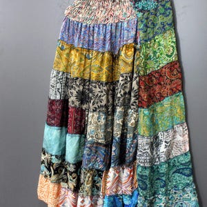 May include: A long, patchwork skirt with a tiered design, featuring a variety of colorful fabric patterns. The skirt is hanging on a wooden hanger against a grey background. The skirt's colours include blue, green, yellow, and red.