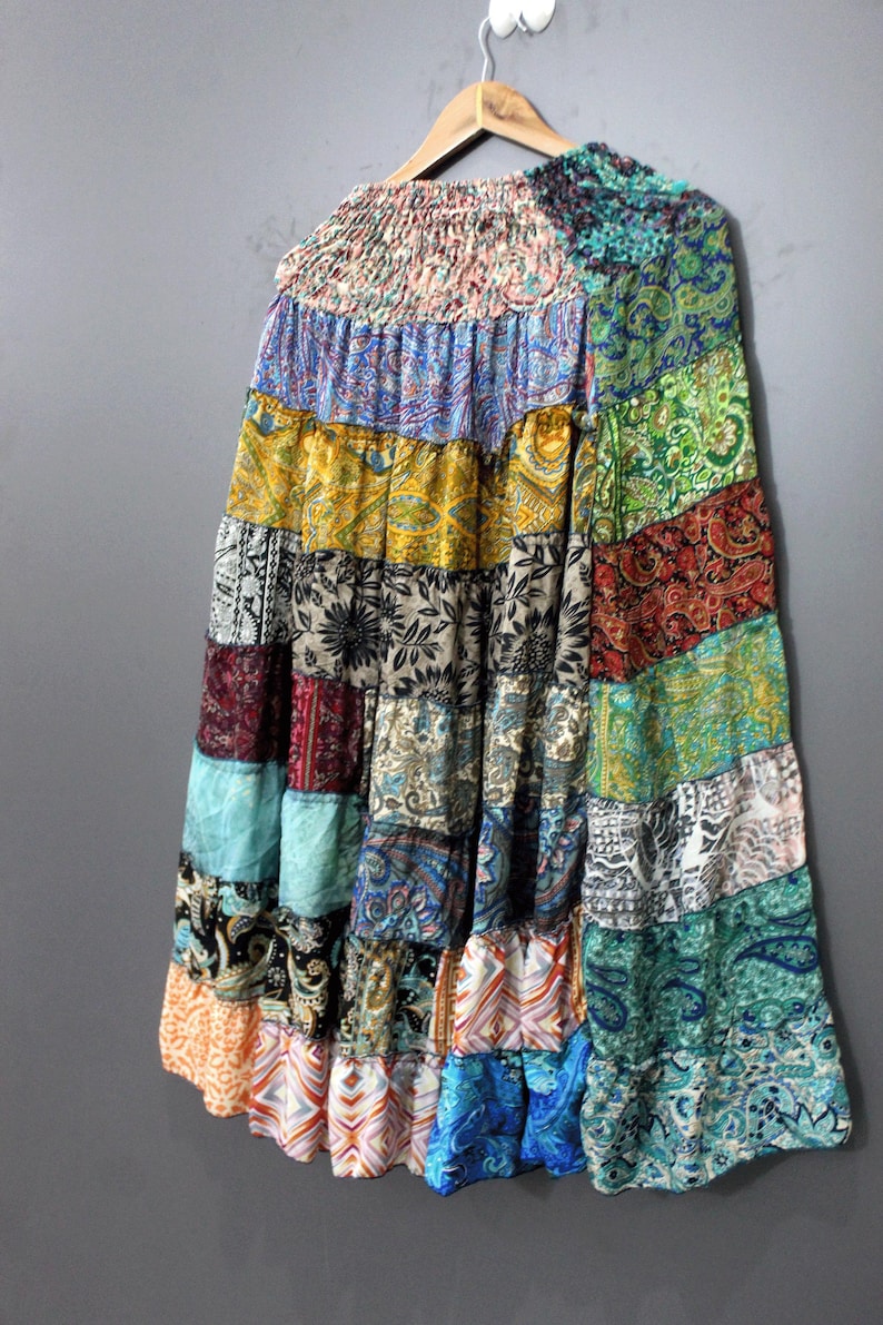 May include: A long, patchwork skirt with a tiered design, featuring a variety of colorful fabric patterns. The skirt is hanging on a wooden hanger against a grey background. The skirt's colours include blue, green, yellow, and red.