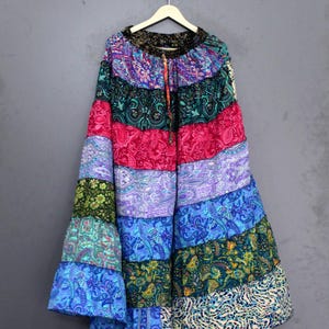 Könnte beinhalten: Ein langer, farbenfroher Rock mit Patchwork-Design. Der Rock hat horizontale Streifen in verschiedenen Farben, darunter Rosa, Blau, Grün und Lila, mit Paisley- und Blumenmustern. Er hängt an einem Holzbügel vor grauem Hintergrund.