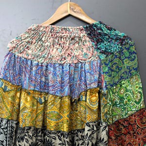 May include: A patchwork skirt in various colours and patterns, including blue, yellow, green, and black. The skirt is hanging on a wooden hanger against a grey background. The skirt has a ruffled design.
