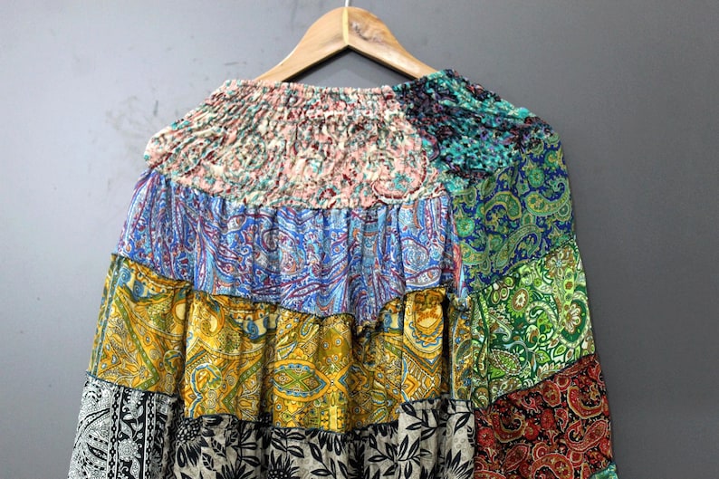 May include: A patchwork skirt in various colours and patterns, including blue, yellow, green, and black. The skirt is hanging on a wooden hanger against a grey background. The skirt has a ruffled design.