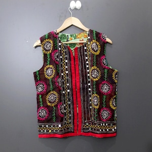 Peut inclure: Un gilet sans manches marron avec broderies florales colorées et des accents miroitants. Le gilet présente une bordure rouge et une doublure à motifs. Le design comprend des broderies jaunes, roses et vertes, créant un vêtement vibrant et accrocheur.
