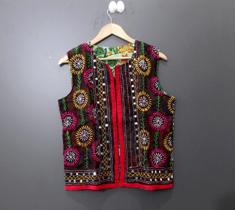 Puede incluir: Un chaleco marr&oacute;n sin mangas con bordados florales coloridos y detalles espejados. El chaleco presenta un ribete rojo y un forro estampado. El dise&ntilde;o incluye bordados amarillos, rosas y verdes, creando una prenda vibrante y llamativa.