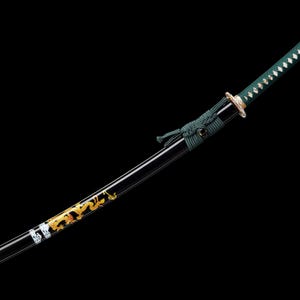 Puede incluir: Una katana japonesa con una vaina negra y un mango verde. La hoja presenta detalles de dragón dorados y blancos. El mango está envuelto en un cordón verde, sobre un fondo negro.