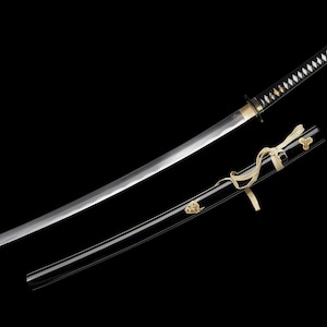 Puede incluir: Una espada katana japonesa con una vaina negra y una hoja plateada. El mango está envuelto en material blanco y negro. La vaina tiene detalles dorados y una cinta color crema. La espada se muestra sobre un fondo negro.