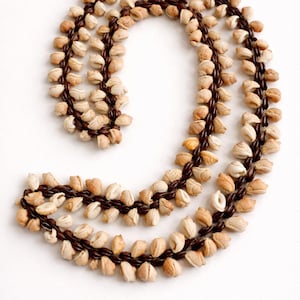 May include: A handcrafted necklace made of small, beige seashells strung together with dark brown cord. The necklace is arranged in a curved, flowing design, showcasing the natural colors and textures of the shells.