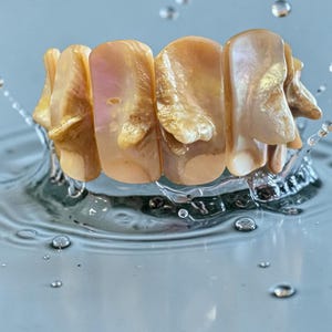 May include: A close-up shot of a bracelet made of large, irregular, light-colored beads. The beads have a pearlescent sheen and are arranged in a row. The bracelet is partially submerged in water, with droplets and ripples visible around it.