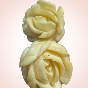 Puede incluir: Dos esculturas de rosas talladas en color marfil. Las rosas están en plena floración, con pétalos detallados. El fondo es un degradado rosa suave. Las esculturas son probablemente artículos decorativos o componentes de joyería.