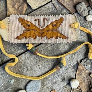 Pode incluir: Pulseira artesanal com contas com um desenho de borboleta em tons de dourado e castanho. A pulseira apresenta uma base de contas de cor creme e é presa a uma tira de couro amarela. A pulseira tem cerca de 3,8 cm de largura.