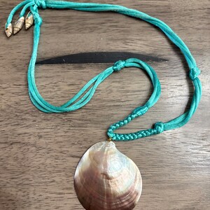 May include: A necklace featuring a large, iridescent, brown and white seashell pendant suspended from a bright turquoise cord. The cord includes a braided section, several knots, and three small, pointed spiral shells at the end of the ties.