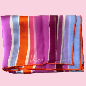 May include: Folded silk scarf with vertical stripes in shades of purple, orange, blue, and white. The scarf has an orange border and the signature "Vana" in white. The scarf is a fashion accessory.