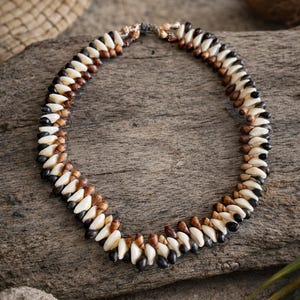 May include: A handcrafted necklace composed of alternating white, brown, and black beads. The beads are arranged in a circular pattern, creating a unique and natural aesthetic. The necklace rests on a weathered wooden surface.