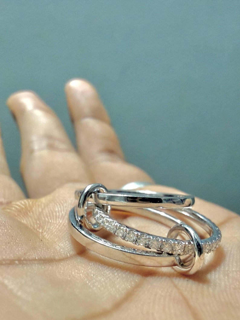 Pu&ograve; includere: Un anello color argento dal design multistrato composto da tre fasce circolari interconnesse. Una delle fasce &egrave; incastonata con una fila di piccole pietre chiare e scintillanti, mentre le altre due fasce sono lisce e lucide.