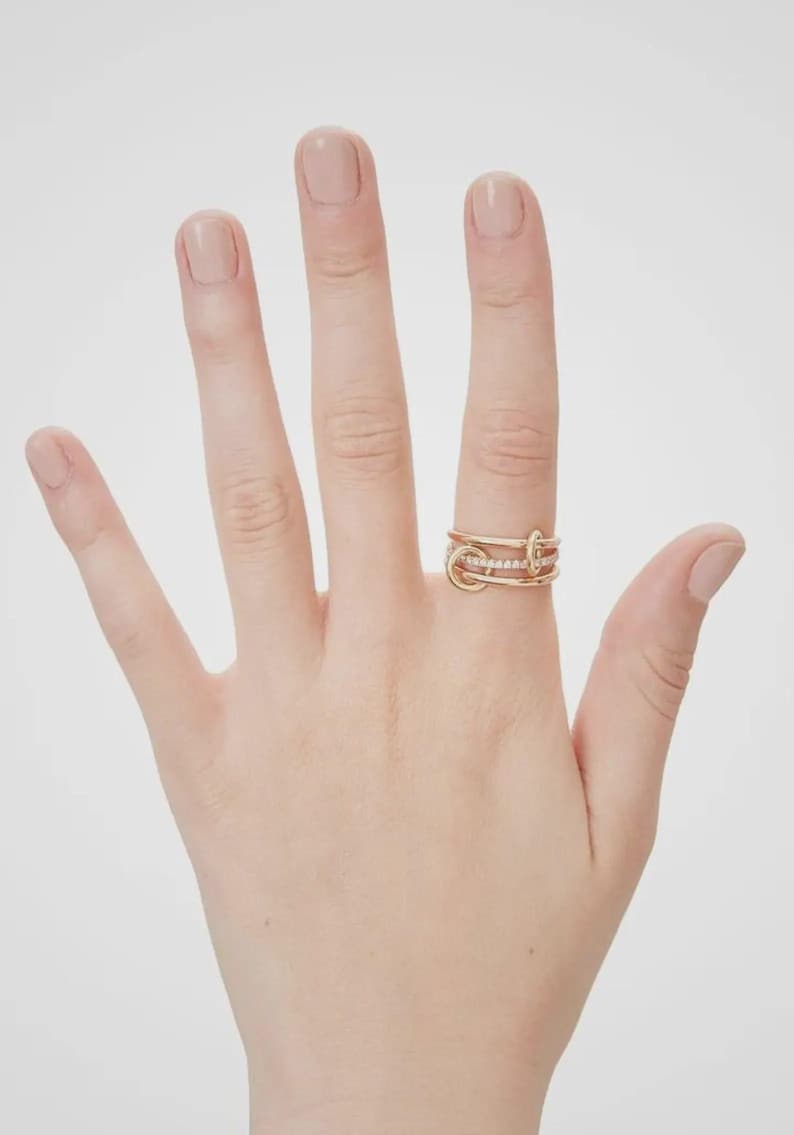 Pu&ograve; includere: Un anello multi-fascia color oro caratterizzato da tre cerchi interconnessi. La fascia centrale &egrave; incastonata con una fila di piccole pietre chiare e scintillanti, mentre le fasce superiore e inferiore sono realizzate in metallo liscio e lucido.