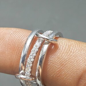 Pu&ograve; includere: Un anello multi-fascia color argento composto da tre bande distinte collegate da due barre orizzontali. La fascia centrale &egrave; incastonata con una fila di piccole pietre chiare e scintillanti, mentre le due fasce esterne hanno una finitura liscia.