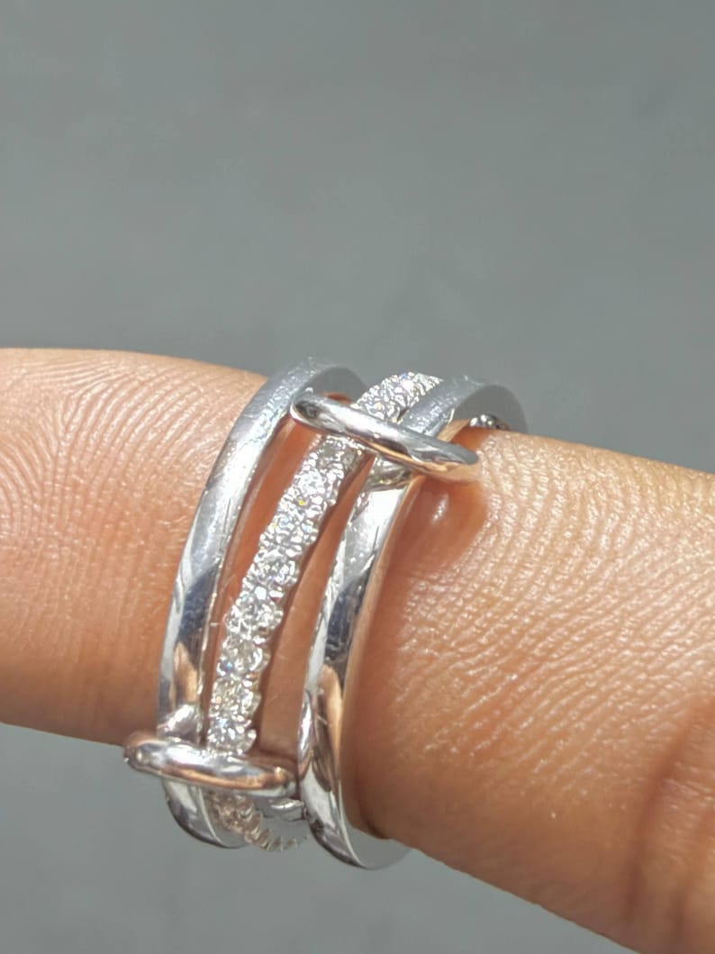 Pu&ograve; includere: Un anello multi-fascia color argento composto da tre bande distinte collegate da due barre orizzontali. La fascia centrale &egrave; incastonata con una fila di piccole pietre chiare e scintillanti, mentre le due fasce esterne hanno una finitura liscia.