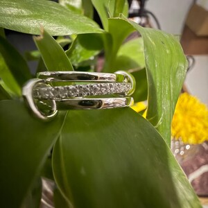 Pu&ograve; includere: Un anello color argento composto da tre fasce distinte. La fascia centrale &egrave; incastonata con una fila di piccole pietre chiare e scintillanti, mentre le due fasce esterne sono lisce, lucide e semplici, creando un aspetto a strati.