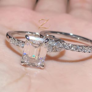 Puede incluir: Dos anillos de plata con piedras preciosas transparentes. Un anillo presenta una piedra central grande de corte rectangular, mientras que el otro tiene una banda adornada con piedras redondas más pequeñas. Los anillos están sobre un fondo borroso.