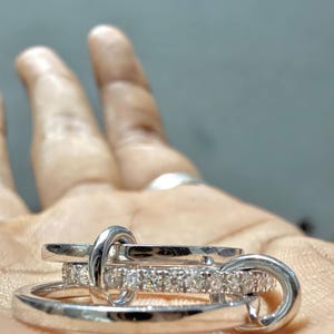 Pu&ograve; includere: Un set di anelli color argento composto da tre fasce intrecciate. Una fascia presenta una fila di piccole pietre chiare e scintillanti sulla superficie esterna, mentre le altre due sono lisce, lucide e realizzate in metallo color argento.