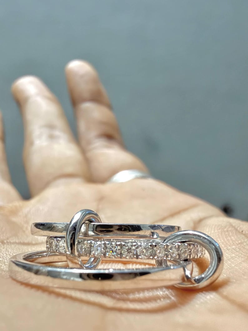 Pu&ograve; includere: Un set di anelli color argento composto da tre fasce intrecciate. Una fascia presenta una fila di piccole pietre chiare e scintillanti sulla superficie esterna, mentre le altre due sono lisce, lucide e realizzate in metallo color argento.