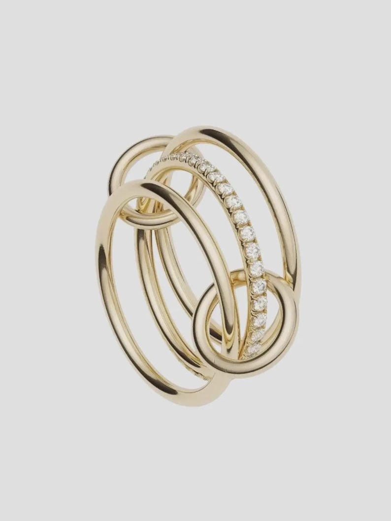 Pu&ograve; includere: Un anello a pi&ugrave; fasce realizzato in oro giallo lucido, caratterizzato da tre cerchi intrecciati. Una delle fasce &egrave; incastonata con una fila continua di piccoli diamanti bianchi rotondi e chiari che catturano la luce, creando un effetto a strati.
