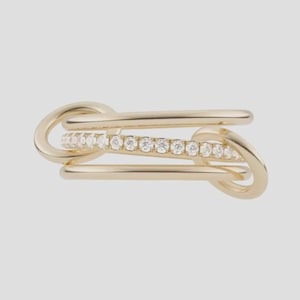 May include: A gold ring featuring a multi-band design with two smooth, polished gold bands flanking a central band embedded with a row of small, clear, round-cut stones. The bands are connected by two circular gold loops on each side.