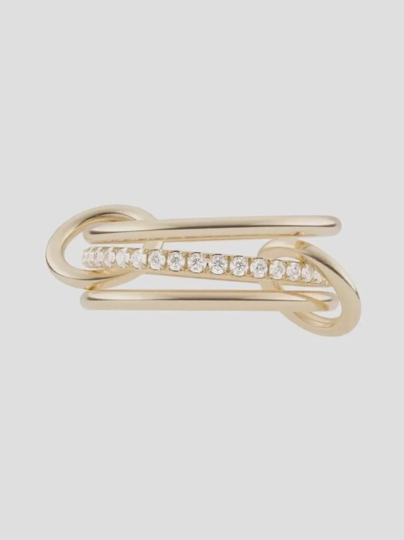 Pu&ograve; includere: Un anello in oro dal design a fasce multiple, composto da due fasce lisce e lucide che ne fiancheggiano una centrale, incastonata con una fila di piccole pietre chiare a taglio rotondo. Le fasce sono collegate da due anelli circolari in oro.