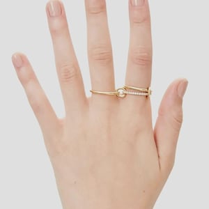 Pu&ograve; includere: Un anello in oro con un design a doppio cerchio collegato da un piccolo anello circolare. Un cerchio &egrave; in oro lucido liscio, mentre l'altro &egrave; incastonato con una fila di piccole pietre chiare e scintillanti sulla superficie anteriore.
