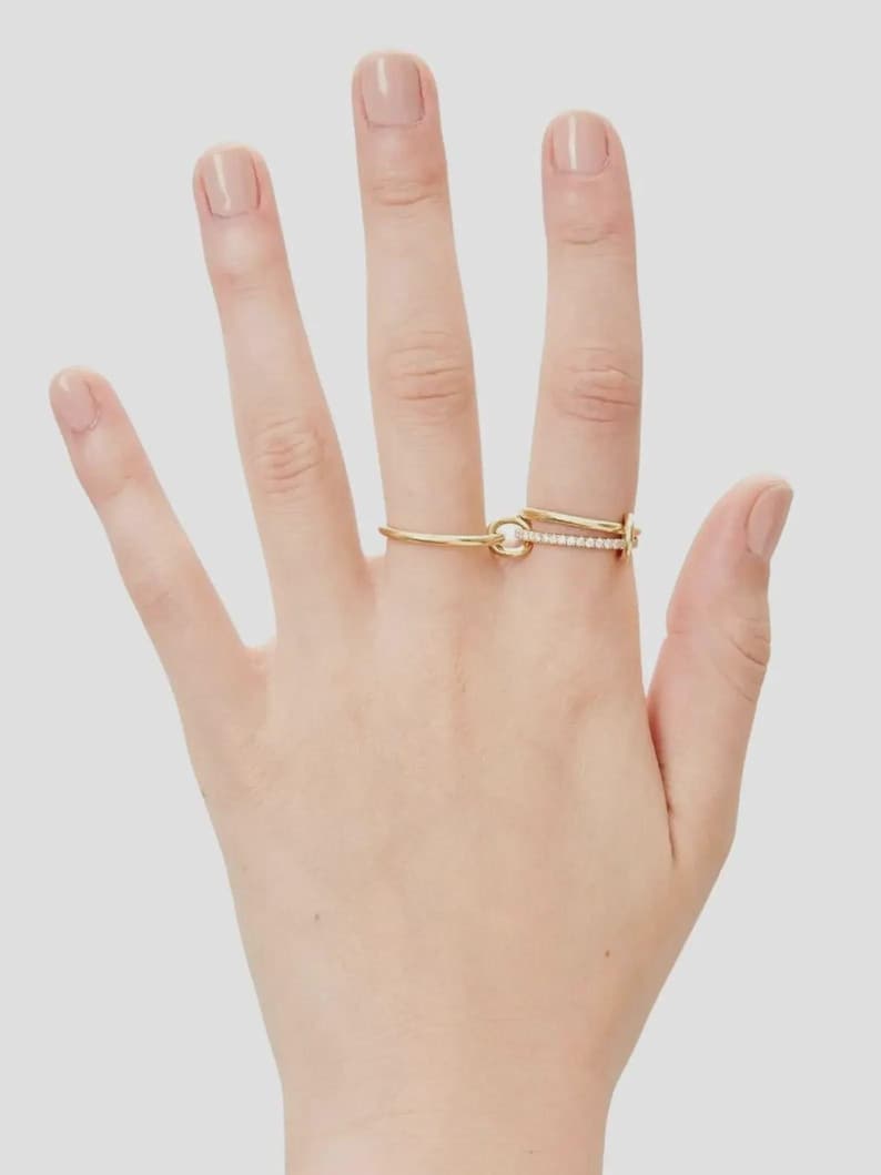 Pu&ograve; includere: Un anello in oro con un design a doppio cerchio collegato da un piccolo anello circolare. Un cerchio &egrave; in oro lucido liscio, mentre l'altro &egrave; incastonato con una fila di piccole pietre chiare e scintillanti sulla superficie anteriore.