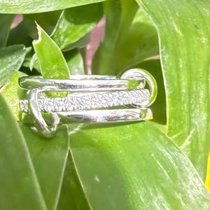 Pu&ograve; includere: Un anello multi-fascia color argento composto da una combinazione di fasce in metallo liscio e lucido e una fascia centrale con una finitura testurizzata e scintillante. Il design presenta diversi anelli intrecciati tra loro.