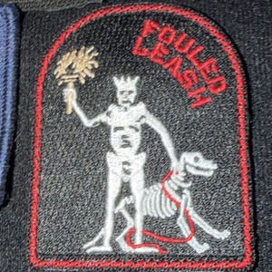 Könnte beinhalten: Eine Sammlung gestickter Aufnäher, darunter eine amerikanische Flagge, ein Streifendesign und ein schwarzer Aufnäher mit einer Skelettfigur, die einen Stab hält und einen Skeletthund an der Leine führt. Der Text "FOULED LEASH" ist über der Figur angeordnet.